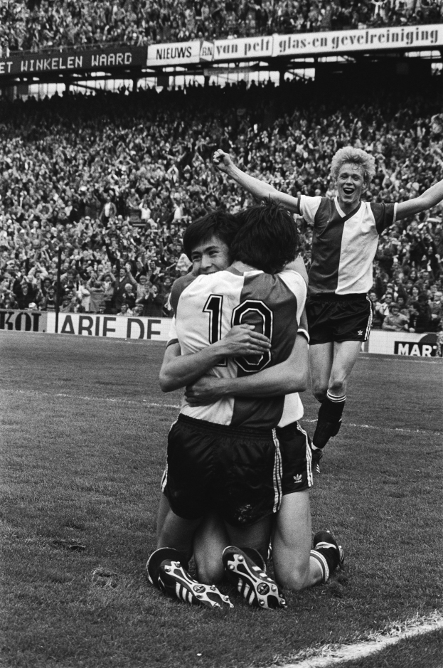 Drei Fußballspieler umarmen sich auf dem Boden nach einem Tor, während eine große jubelnde Menge Schilder schwenkt in einem schwarz-weißen Stadion.