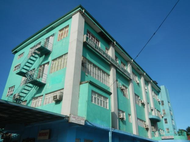 Ein großes grünes Gebäude mit zahlreichen Fenstern, Klimaanlagen, Treppen, Geländern, Schilden, Strommasten, Kabeln, Fahrzeugen auf der Straße, Bäumen und einem klaren blauen Himmel.