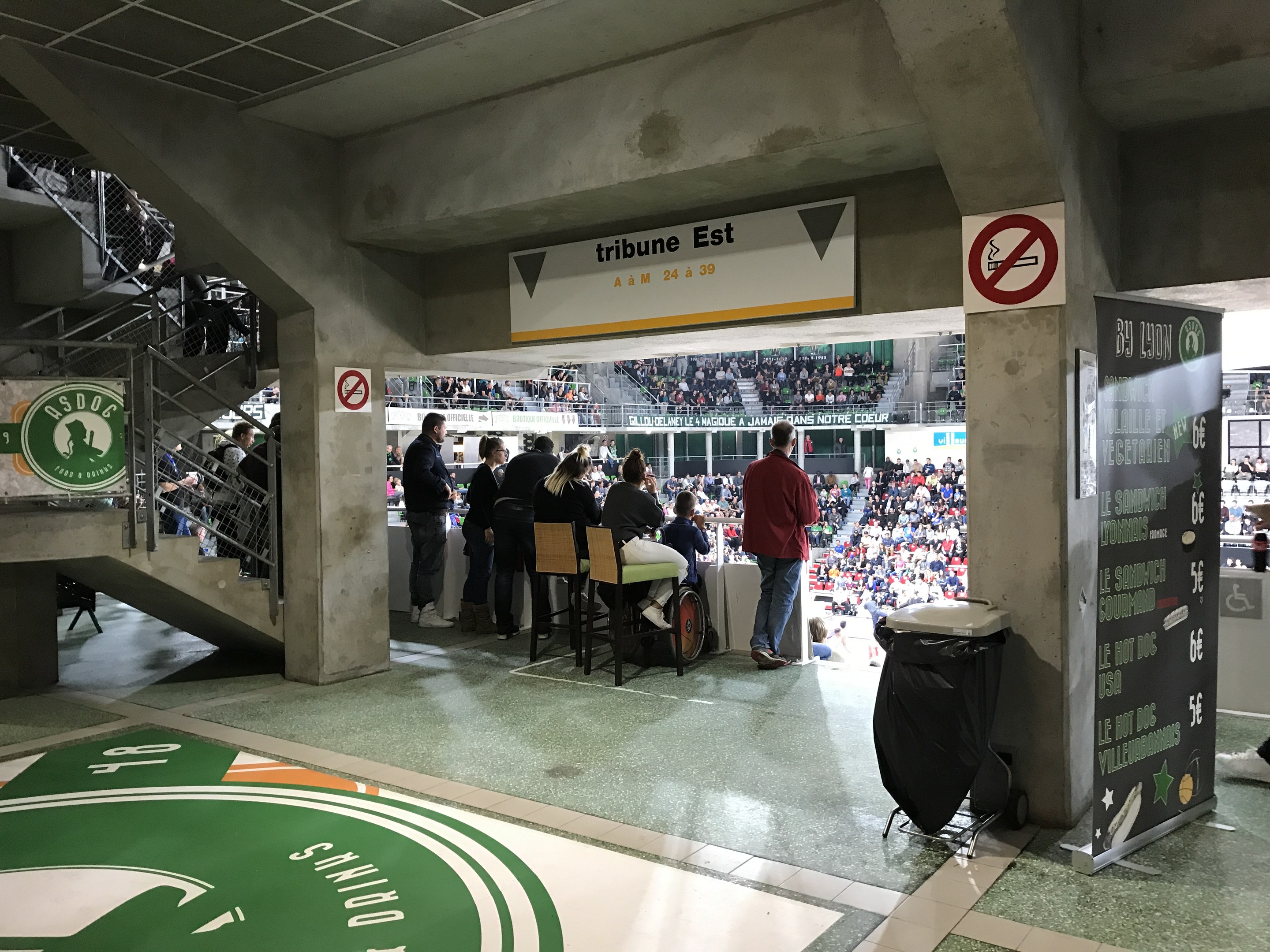 Gruppe von Menschen, die vor einer sitzenden Menge in einem Stadion mit Schildern, einem Müllcontainer, einer Tafel, einer Treppe und einem Dach mit Lampen stehen, mit der Aufschrift "Boston Celtics vs New York Jets" auf den Schildern.