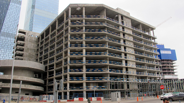 Großes Bauprojekt in einer städtischen Umgebung mit Hochhäusern, Fahrzeugen, Fußgängern, Verkehrsinfrastruktur und Kränen vor einem klaren Himmel.