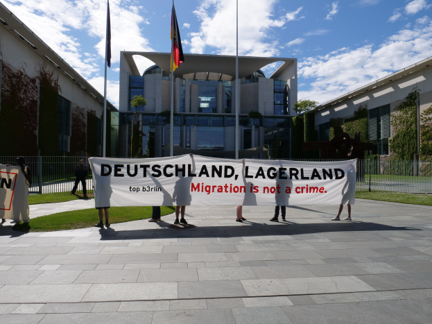 Eine Gruppe von Menschen hält ein Transparent mit der Aufschrift "Deutschland, Lagerland Migration ist kein Verbrechen" vor einem umzäunten Gebäude mit Bäumen und Fahnen.