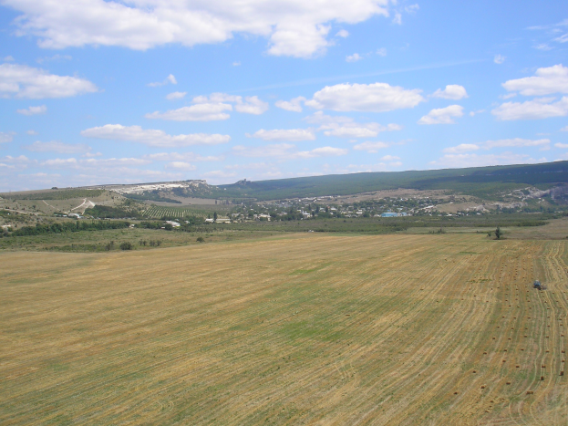 Ein Traktor steht inmitten eines bewirtschafteten Feldes, umgeben von Bäumen, Gebäuden, Hügeln und einem bewölkten Himmel.