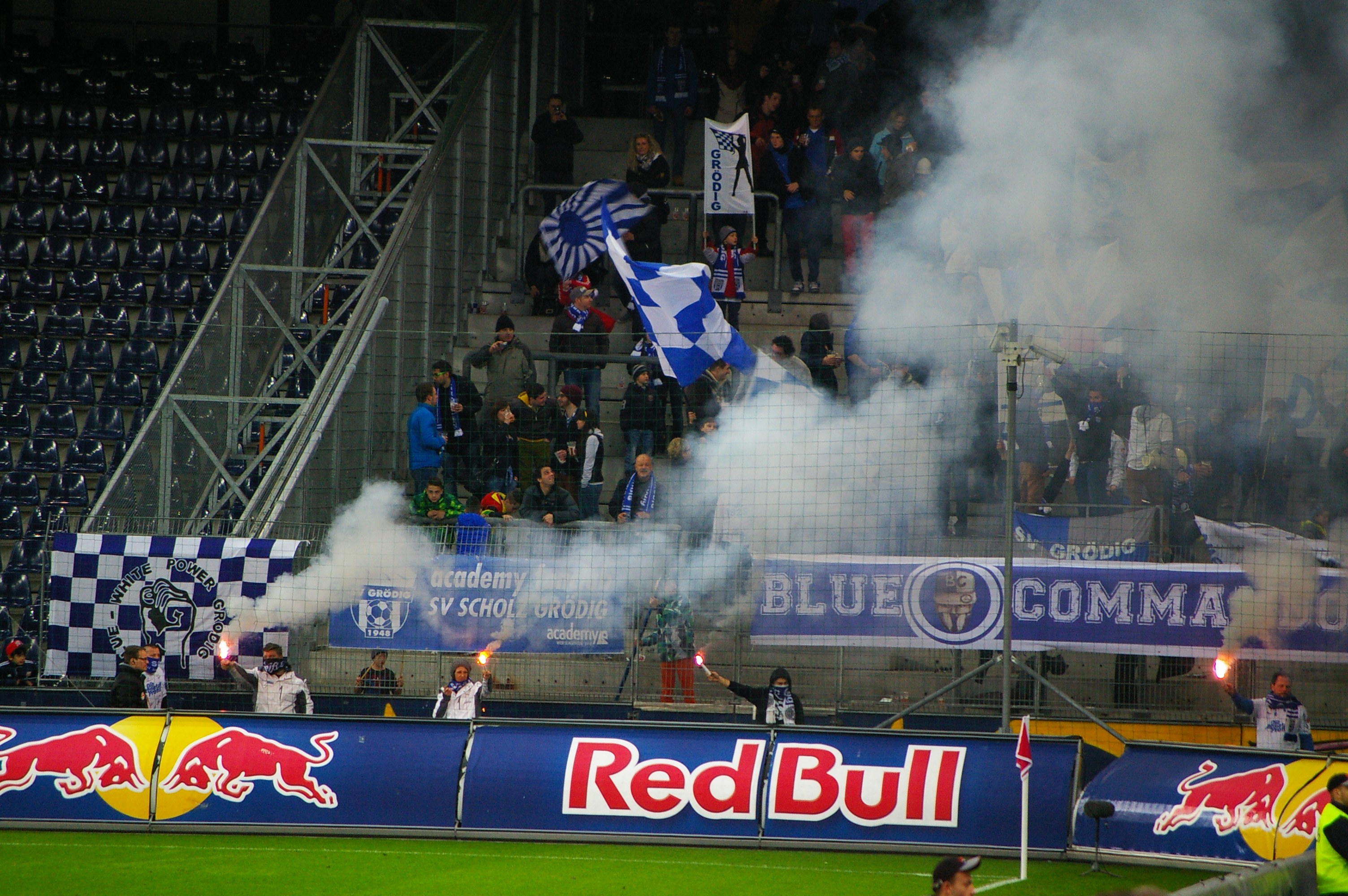 Eine Gruppe von Menschen auf einem Fußballfeld, die Fahnen und Banner halten und Rauch von der Erde aufsteigt, umgeben von einem Metallzaun und leeren Stadionstühlen.