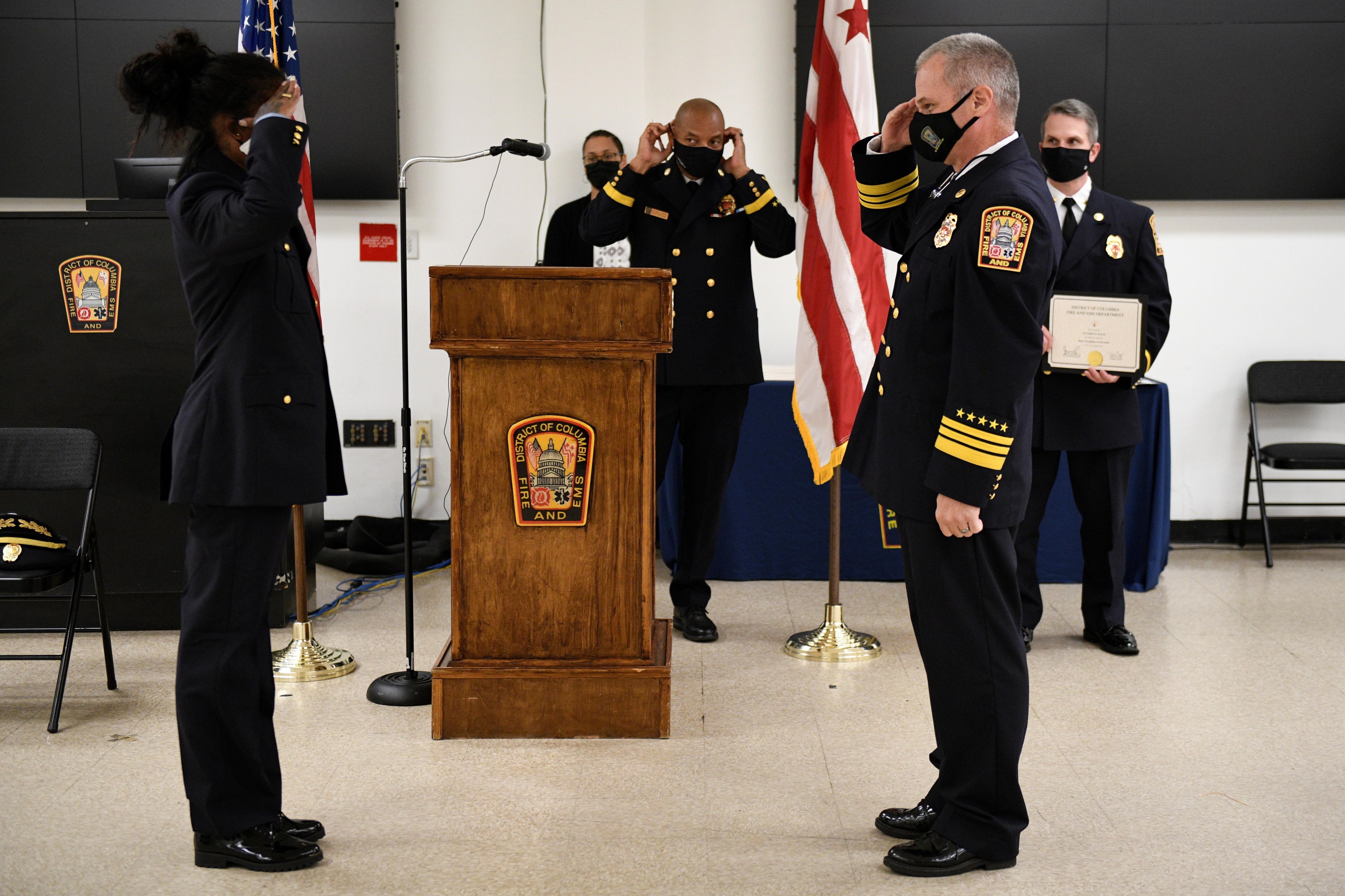 Gruppe uniformierter Personen in Masken vor einem Rednerpult mit Mikrofon während einer Vereidigung eines neuen Feuerwehrchefs, der eine Urkunde hält und von Fahnen, Stühlen, Tischen und einer Wand umgeben ist.