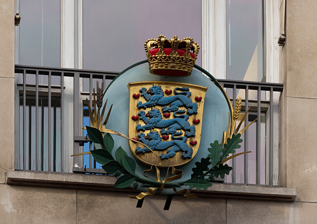 Das Wappen der britischen Königsfamilie auf der Außenseite eines Gebäudes dargestellt, begleitet von einem Kronen- und Blätter-Logo, mit Fenstern und Metallgittern im Hintergrund.