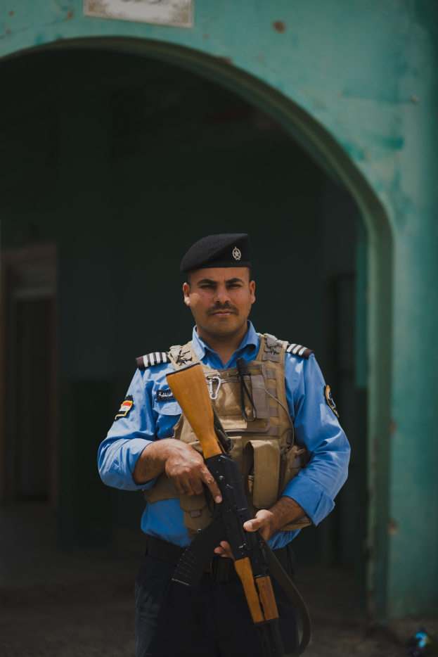 Polizist in Uniform mit Gewehr vor einem Gebäude mit einem Torbogen und einer Tafel an der Wand.