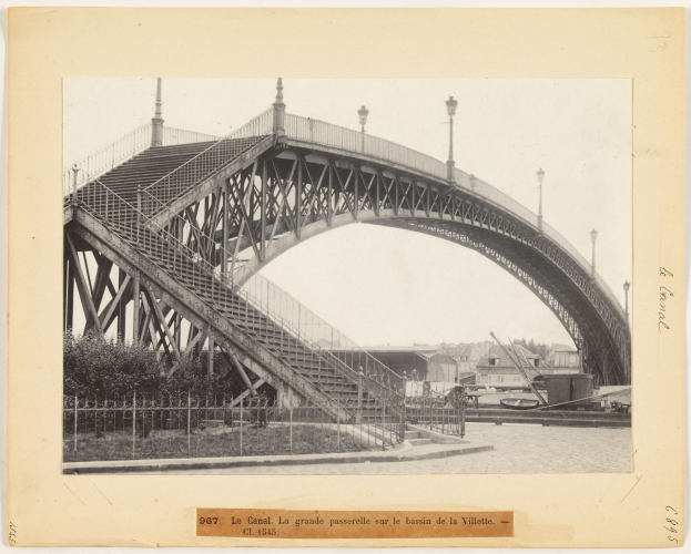 Schwarzes und weißes Foto einer Brücke mit Treppen, Geländern, Pfosten, Lampen, Bäumen und Gebäuden im Hintergrund, mit Text unten.