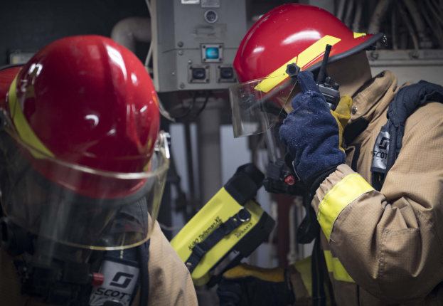 Zwei Feuerwehrleute in Schutzausrütstück arbeiten an einem Feuerhydranten während einer Ausbildung mit Maschinen und Kabeln im Hintergrund.