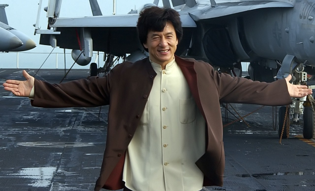 Jackie Chan steht auf dem Deck eines Flugzeugträgers mit ausgestreckten Armen in einem weißen und braunen Kleid, während Kampfjets und ein klarer blauer Himmel im Hintergrund zu sehen sind.