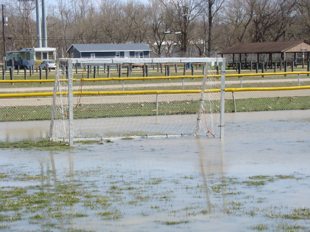 Ein Fußballtor steht in der Mitte eines überfluteten Feldes, umgeben von Gras und Wasser, sowie Schuppen, Pfosten, Bäumen, Fahrzeugen und einem klaren blauen Himmel im Hintergrund.