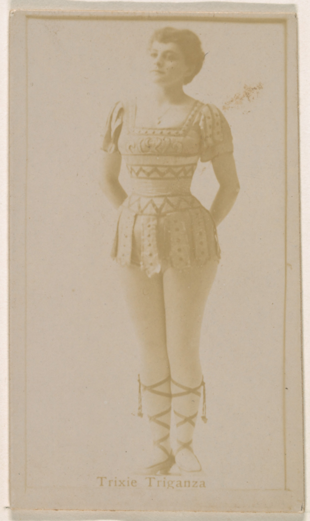 Ein altes Foto einer Frau in einem hellfarbenen gemusterten Badeanzug, die Haare zu einem Knoten gebunden, steht und lächelt direkt in die Kamera mit ihren Händen vor der Brust verschränkt.