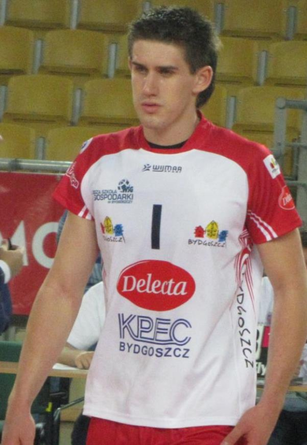 Ein Mann in rot-weißer Handballdress steht auf einem Feld, umgeben von Zuschauern, mit einer roten Fahne im Hintergrund, auf der "Krasnoyarsk vs Bydgoszcz" steht.