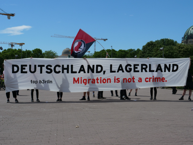 Eine Gruppe von Menschen mit einem Transparent, auf dem "Deutschland, Lagerland Migration ist kein Verbrechen" steht, und einer Flagge, im Hintergrund Straßenlaternen, Bäume, Kräne, ein Gebäude und ein bewölkter Himmel.