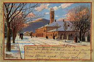 Ein altes Postkartenbild, das ein schneebedecktes Gebäude umgeben von Bäumen, Menschen und einem Zaun zeigt, mit der Aufschrift "Freiburg Hauptbahnhof" unten.