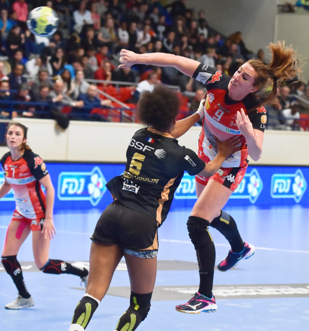 Eine Gruppe von Frauen spielt Handball auf einem Platz mit einem Ball in der Luft, beobachtet von Zuschauern in einer Stadionatmosphäre.