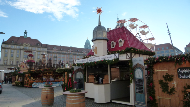 Ein belebter Weihnachtsmarkt auf einem Stadtplatz mit geschmückten Bäumen, Gebäuden, Fahrzeugen, Passanten und wolkenverhangenem Himmel.