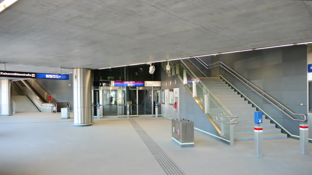 Leerer Bahnhof mit Treppe, Geländern, bodennahen Mülltonnen, wandmontierten Texttafeln, Deckenlampen und Glastüren im Hintergrund.