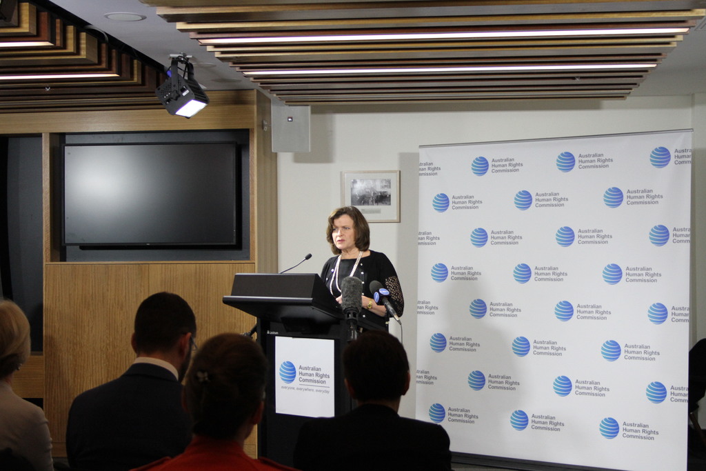 Eine Frau steht auf einer Bühne und spricht in ein Mikrofon, vor ihr sitzt ein Publikum, hinter ihr hängt ein Banner an der Wand, ein Fernseher an der Holzwand und Lampen an der Decke.