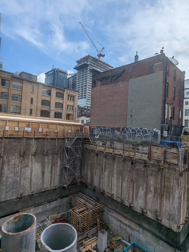 Luftaufnahme einer Baustelle mit freiliegenden Rohren und Stangen darunter, umgeben von Gebäuden mit Fenstern unter einem bewölkten Himmel.