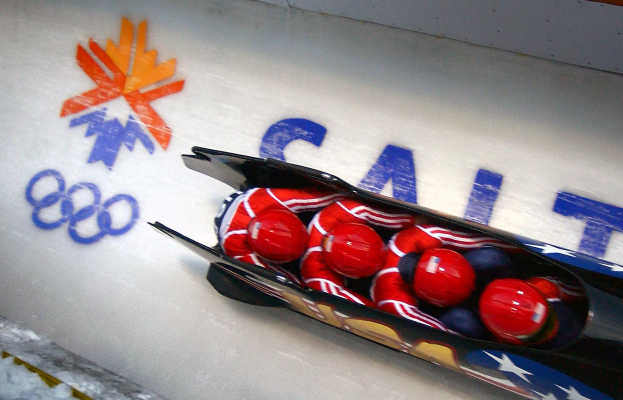 Ein Bobfahrer in Aktion bei den Olympischen Spielen, mit einem Fahrzeug im Vordergrund und einer Wand mit Text und einem Gemälde im Hintergrund.