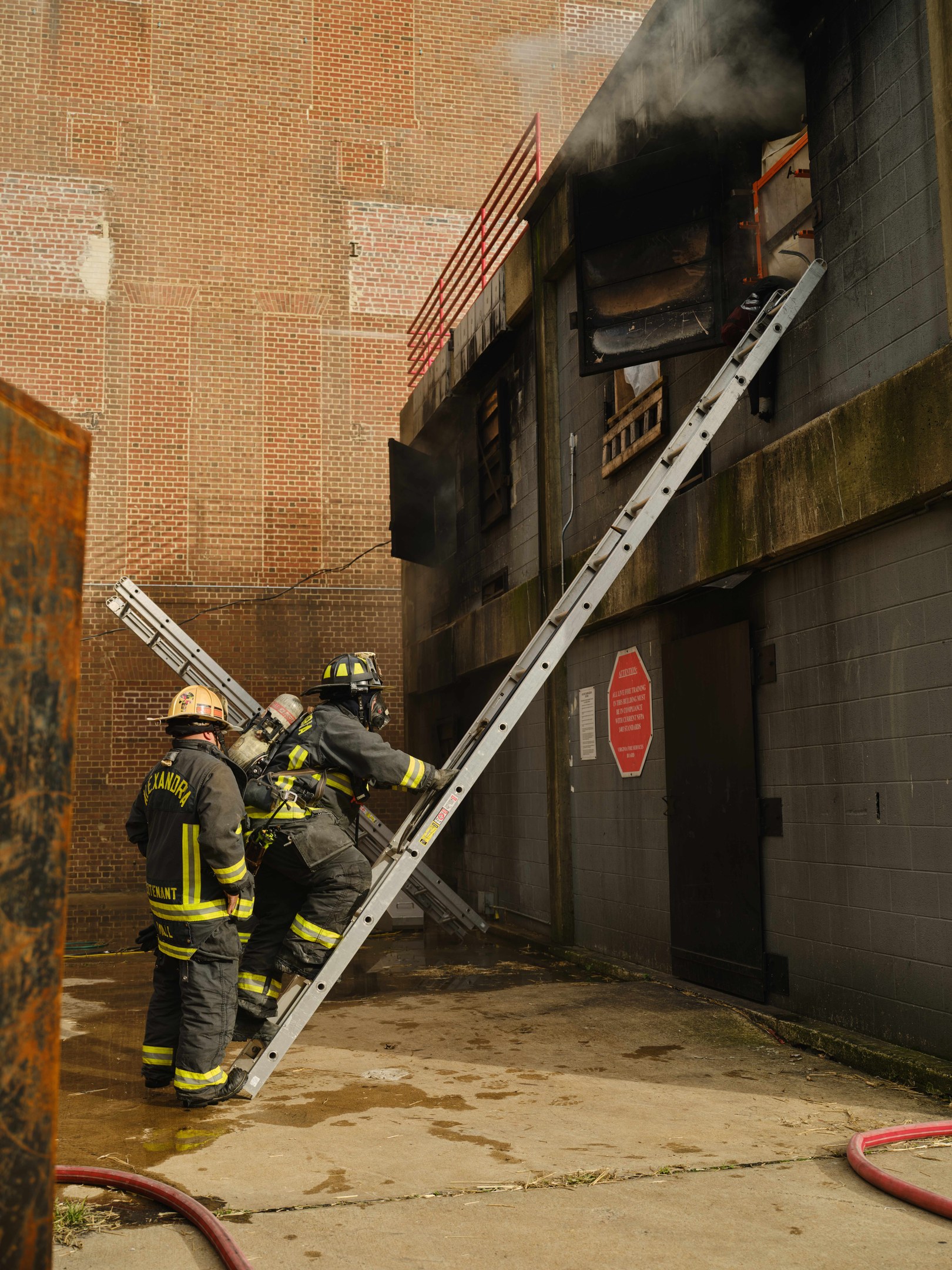 Feuerwehrleute in Helmen klettern eine Leiter zu einem raucherfüllten Gebäude hinauf, mit einem Metallobjekt links und sichtbaren Rohren am Boden.