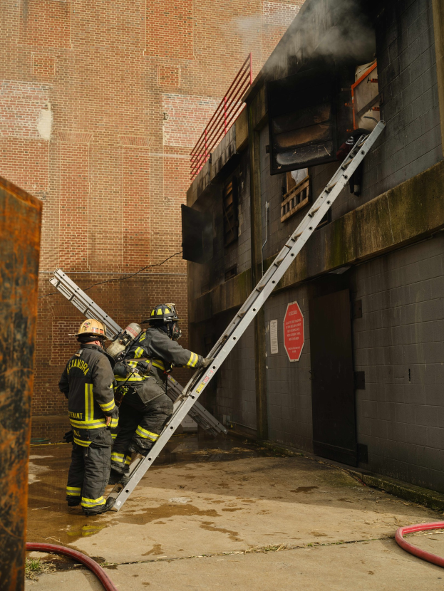 Feuerwehrleute in Helmen klettern eine Leiter zu einem raucherfüllten Gebäude hinauf, mit einem Metallobjekt links und sichtbaren Rohren am Boden.