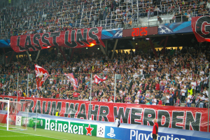 Ein Fußballspiel wird in einem Stadion mit einer großen Zuschauermenge, grünem Rasen, einem Torpfosten links, Bannern auf dem Boden und Menschen mit Fahnen in der Hand gespielt. Im Hintergrund sind Geländer, Tafeln mit Text, Lampen und andere Gegenstände zu sehen.