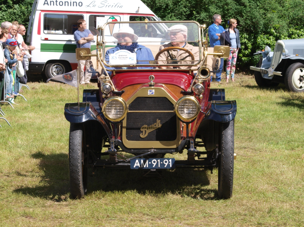 Eine Gruppe von Menschen, die um ein Oldtimer-Auto in einem Feld stehen, mit zwei Personen im Auto sitzend; Stühle sind links zu sehen, und Bäume und ein klarer blauer Himmel bilden den Hintergrund.