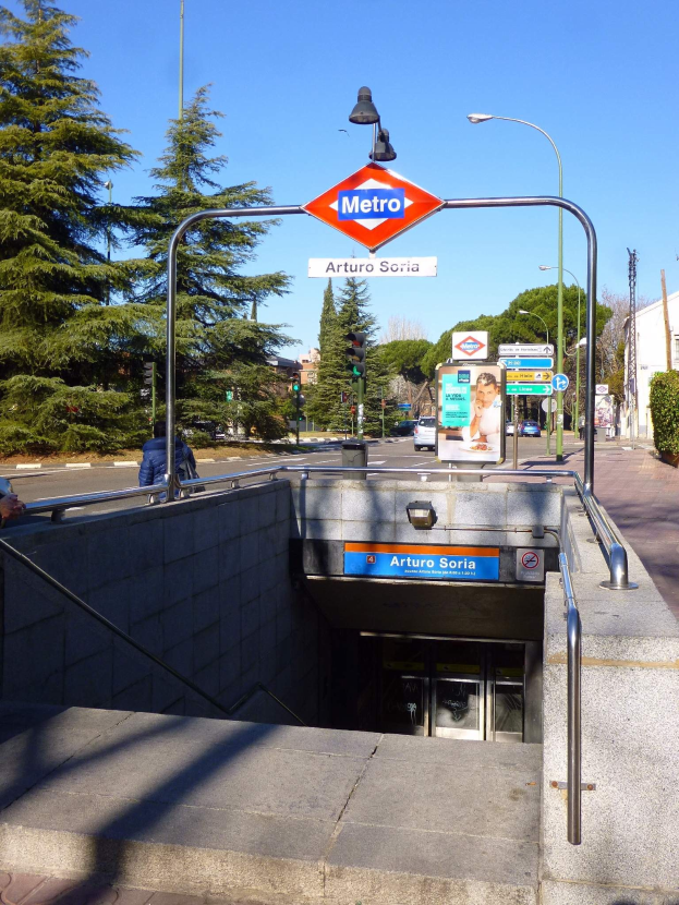 Eine belebte Stadtstraße mit Fahrzeugen, Fußgängern, Gebäuden, Bäumen und einer "Metro"-Anzeige unter einem bewölkten Himmel.