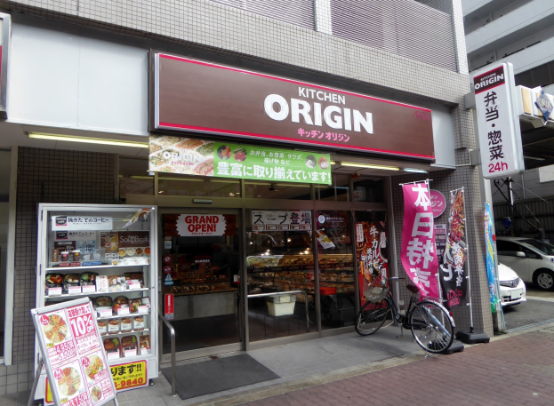 Ladenfront mit Glaswänden und -türen, mit einem geparkten Fahrrad davor, Lebensmitteln auf Regalen drinnen und Texttafeln, Bannern; Fahrzeuge auf der Straße rechts sichtbar.