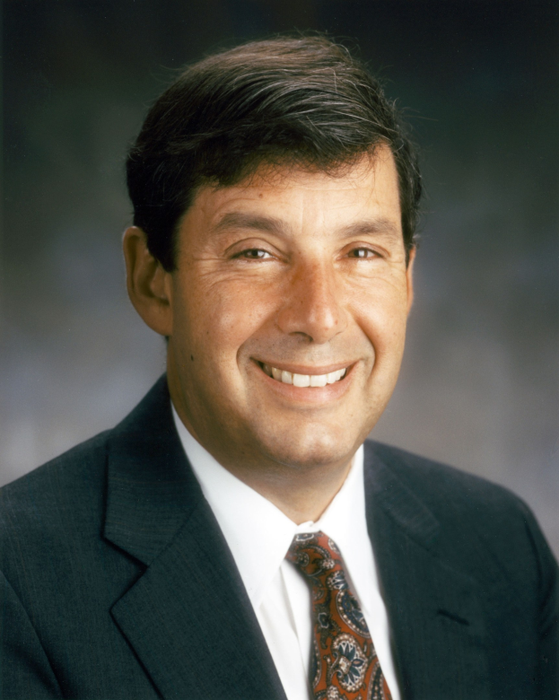 Ein Mann im Anzug und mit Krawatte lächelt in die Kamera und sieht aus wie ein freundlicher Versicherungsmakler vor einem unscharfen Hintergrund.