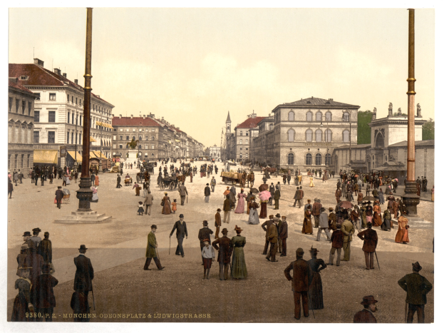 Eine belebte Münchner Stadtstraße aus den frühen 1900er-Jahren mit Menschen, die spazieren gehen, einige mit Schirmen, und einem Pferdewagen im Vordergrund, Gebäuden, Bäumen und Pfählen im Hintergrund und Text am unteren Bildrand.