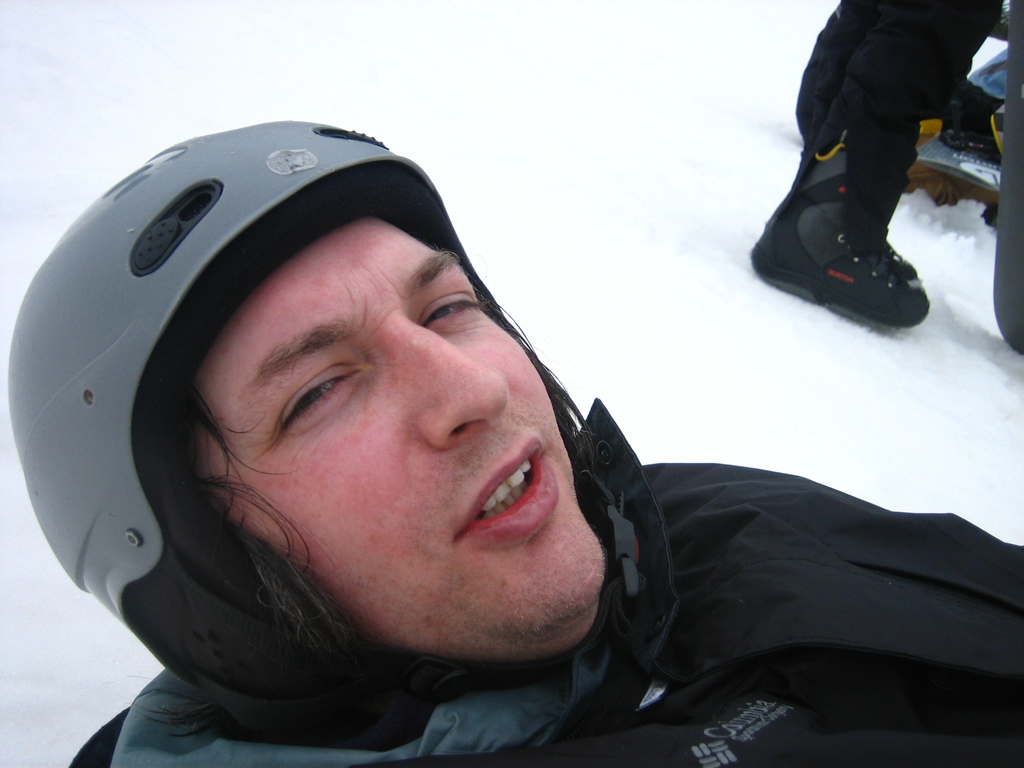 Mann in Pullover und Helm im Schnee, mit den Beinen und Skiern einer anderen Person oben rechts.