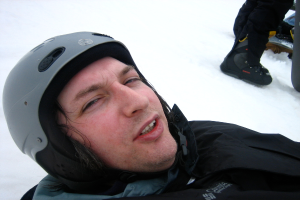 Mann in Pullover und Helm im Schnee, mit den Beinen und Skiern einer anderen Person oben rechts.