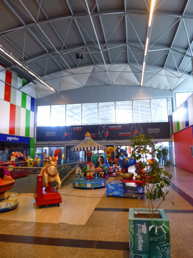 Amusement rides, a potted plant, scattered toys, informational boards, walls, ceiling lights, and metal poles fill a large indoor shopping mall space.