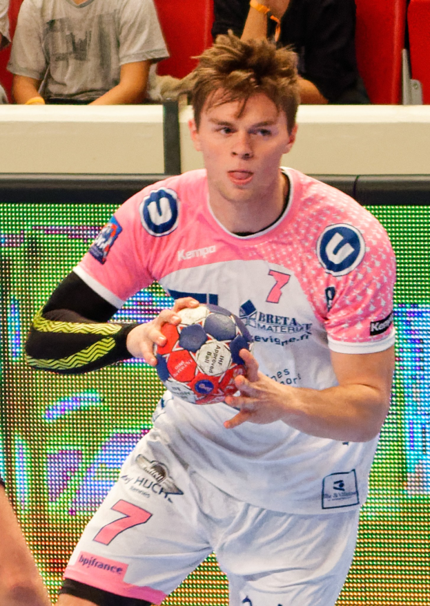 Ein Mann in einem pinken Hemd mit einem Fußball, daneben eine andere Person und im Hintergrund Menschen auf Stühlen sitzend.
