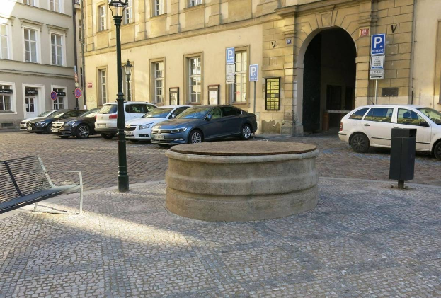 Ein Stadtplatz mit einer Bank, einem Brunnen, Laternen und Schildern, umgeben von Gebäuden mit Fenstern.