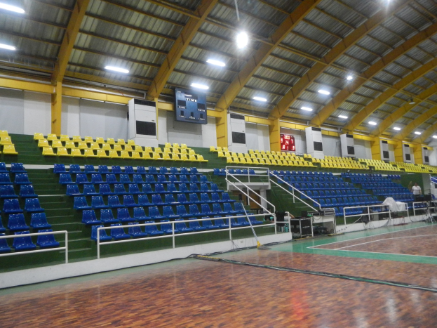 Großer Hallensportplatz mit Holzboden, blauen und gelben Sitzplätzen, Geländern, Treppen mit Geländern, wandmontierten Bildschirmen, Deckenleuchten und einigen Menschen.