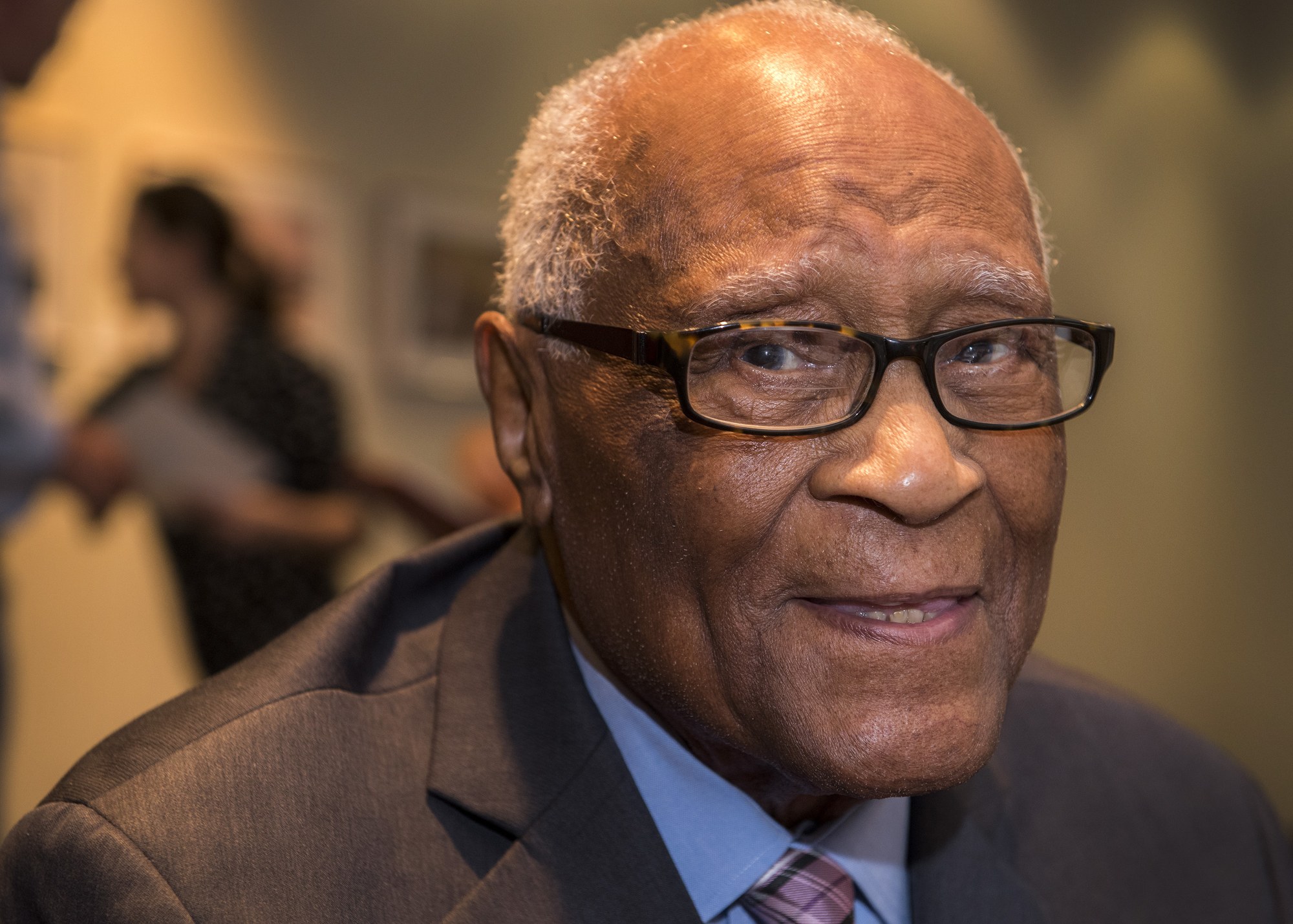 Porträt von Bill Cosby, ein älterer Mann in Anzug und Krawatte, der freundlich lächelt und eine Brille trägt, mit leicht unscharfem Hintergrund.