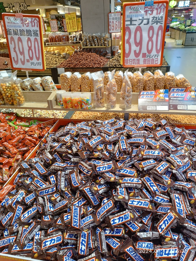 Vitrine mit verschiedenen Süßigkeiten, darunter Snickers-Riegel, in einem Geschäft mit Lebensmitteln auf Regalen und Hintergrunddetails.