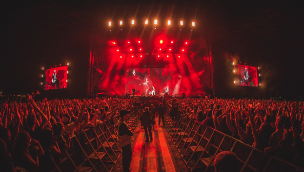 Eine große Menschenmenge steht vor einer hell beleuchteten Bühne mit Geländern, auf der Musiker spielen und andere zusehen, vor einem dunklen Hintergrund.