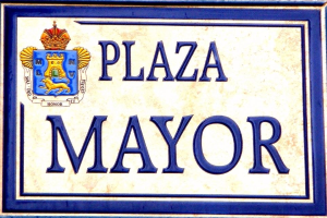 Rechteckige weiße Plakette mit schwarzer Schrift "Plaza Mayor" in fetter Schrift, blauem Rand und einem gekrönten Wappen-Logo an der Seite eines Gebäudes.