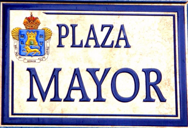 Rechteckige weiße Plakette mit schwarzer Schrift "Plaza Mayor" in fetter Schrift, blauem Rand und einem gekrönten Wappen-Logo an der Seite eines Gebäudes.