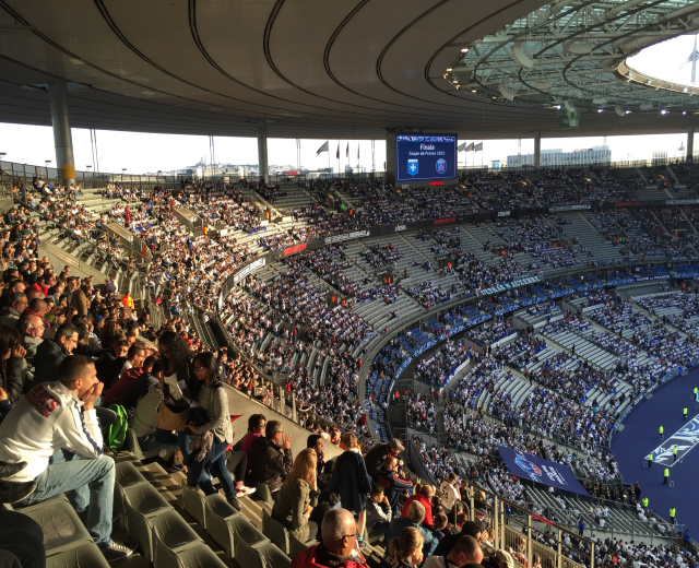 Eine große Menschenmenge in einem Stadion schaut einem Fußballspiel zu, mit einer Bühne rechts, Fahnen, Stangen, einem Bildschirm und der Allianz Arena in München, Deutschland im Hintergrund.