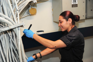 Eine Frau in einem schwarzen Hemd und blauen Handschuhen arbeitet an einem Computer, hält ein Objekt in der Hand und ist von zahlreichen Kabeln auf der linken Seite und einem wandmontierten Kasten im Hintergrund umgeben.