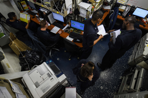 Eine Gruppe von Menschen, die an Büroschreibtischen mit Computern, Tastaturen, Mäusen, Papieren, Büchern und anderen Gegenständen sitzen, umgeben von Maschinen und Papieren, die auf dem Boden verstreut sind, sowie einer textgefüllten Tafel an der Wand.