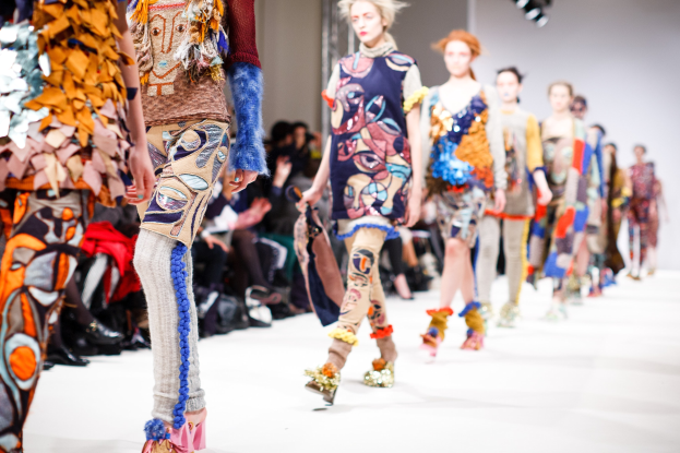 Eine Gruppe Von Frauen Geht Den Laufsteg Bei Der London Fashion Week entlang, Mit Einer Zuschauerschaft Im Hintergrund Unter Hellen Scheinwerfern.
