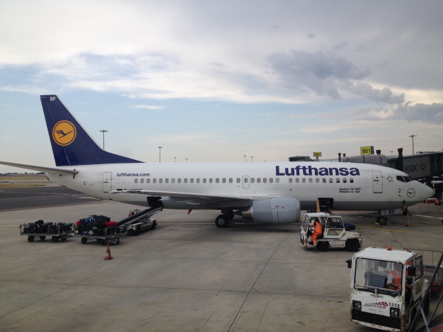 Ein Lufthansa Airbus A320-200 am Frankfurter Flughafen mit Bodenbetriebsfahrzeugen, Verkehrskegeln und Beschilderung im Hintergrund bei bewölktem Himmel.