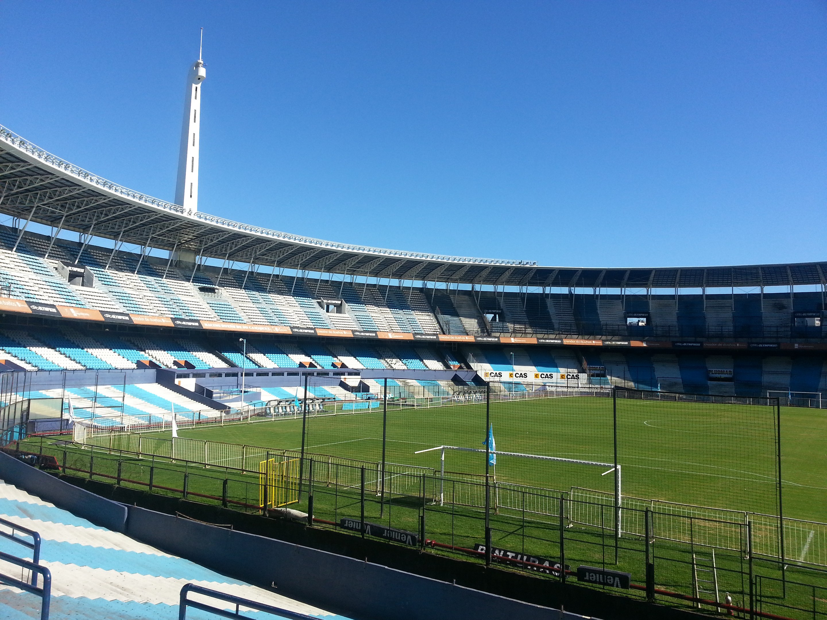 Großes Stadion mit einem Fußballfeld, das von einem Zaun umgeben ist, ein Turm im Hintergrund und ein klarer blauer Himmel.