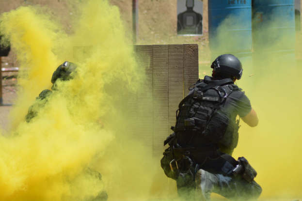 Zwei Polizisten in Einsatzausrüstung atmen gelben Rauch aus, während sie neben einer Wand mit Text auf einer Tafel stehen.